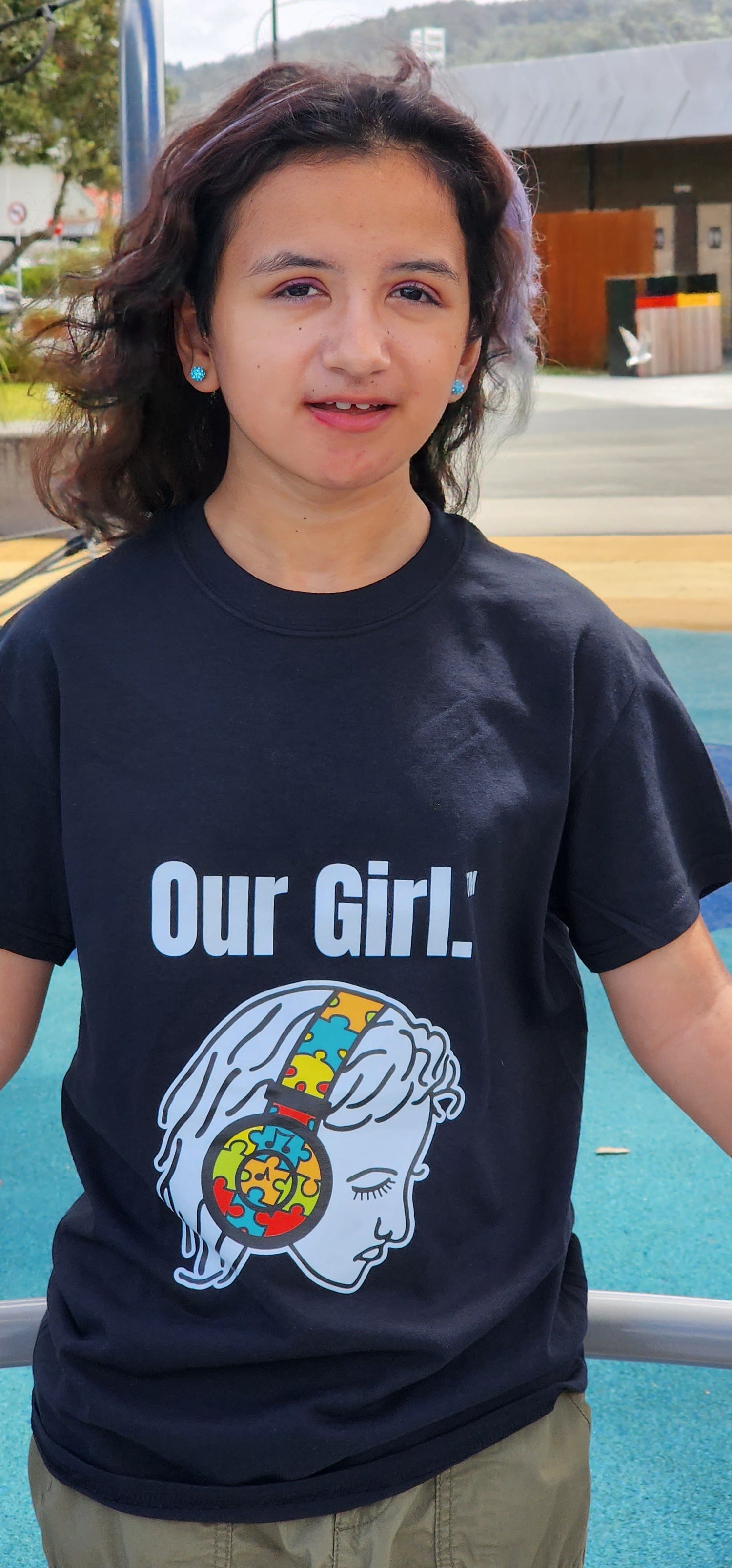 Girl wearing black t-shirt with Our Girl branded on it with a face of a girl wearing headphones with a jigsaw puzzle pattern through-out it.